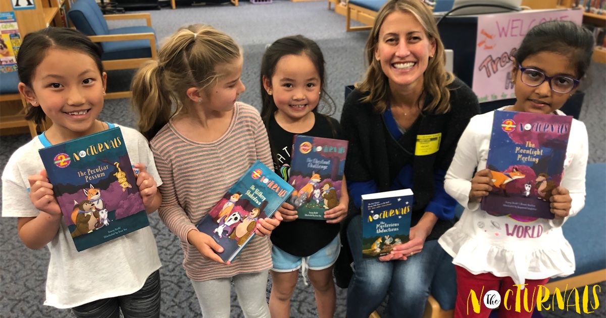 Four girls hold up different titles of The Nocturnals Early Reader series in a school library. Tracey Hecht, the author, is crouched down holding up The Nocturnals: Mysterious Abductions.