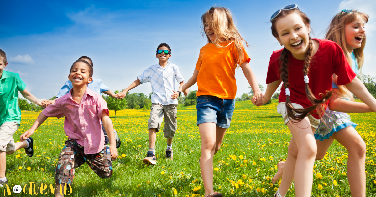 Walking through a flower filled meadow, a group of children are smiling and holding hands while wearing brightly colored shirts. 
