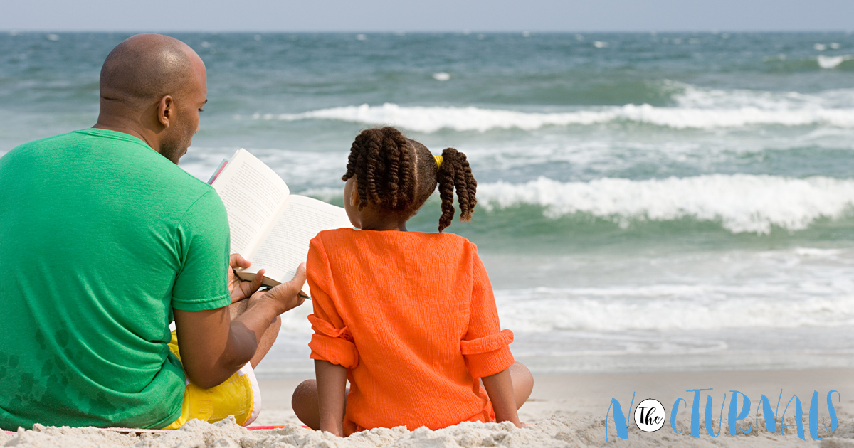 A father is reading to a younger girl while they both sit on the sand in front of the ocean. 