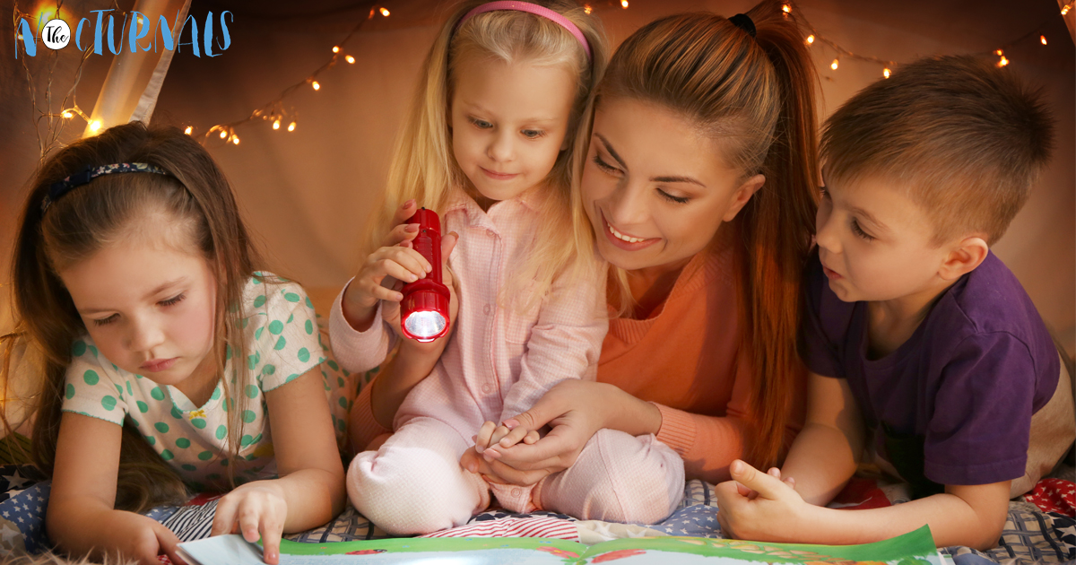 Two girls, a boy, and an older woman are reading a book by flashlight in a tent. 