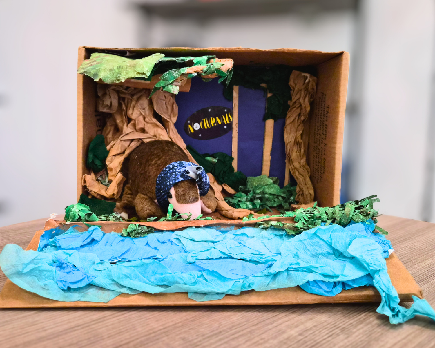 Images shows a completed example of the Nocturnals Habitat Activity. A stuffed pangolin sits in a cardboard box decorated with colorful paper to mimic trees and water.