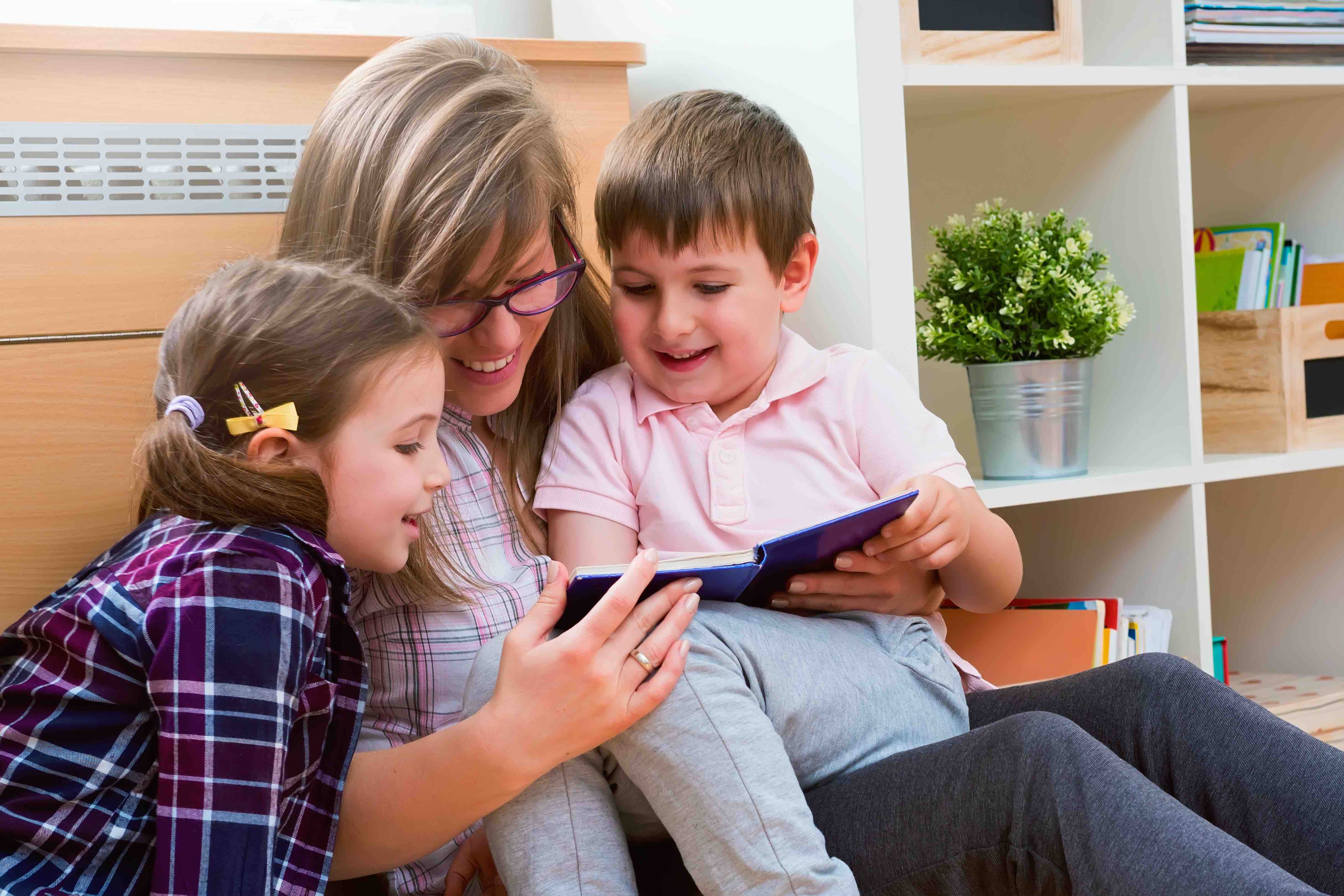 An older woman is sitting down, holding a young boy on her lap, while looking at the book he is holding. Beside the woman is a young girl leaning towards her to see the book. 